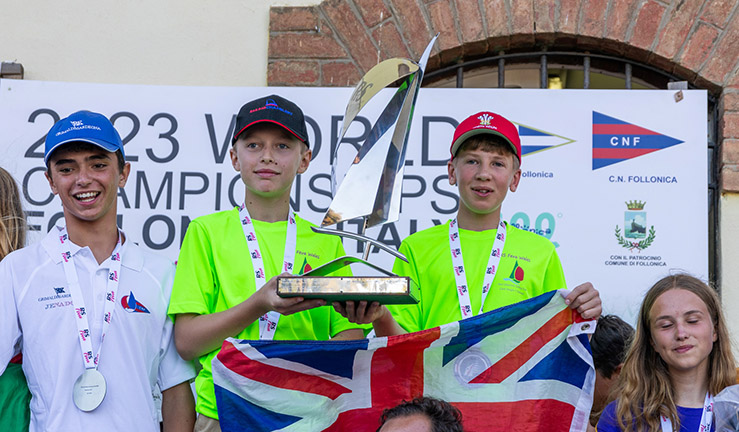 Teenagers Ben Greenhalgh and Tom Sinfield from Port Dinorwic Sailing Club on the podium - with a large boat trophy and Union Jack flag - at the prizegiving after winning the 2023 RS Feva Worlds in Italy.