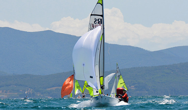 Teenagers Ben Greenhalgh and Tom Sinfield sailing their RS Feva downwind with the kite flying at Follonica, Italy, mountains in the background, at the 2023 RS Feva Worlds in Italy.