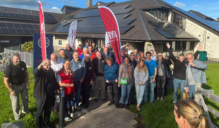 Group of people smiling for the camera outside Plas Menai National Outdoor Centre with RYA feather flags and sunshine for The Big Weekend for clubs, training centres and instructors.