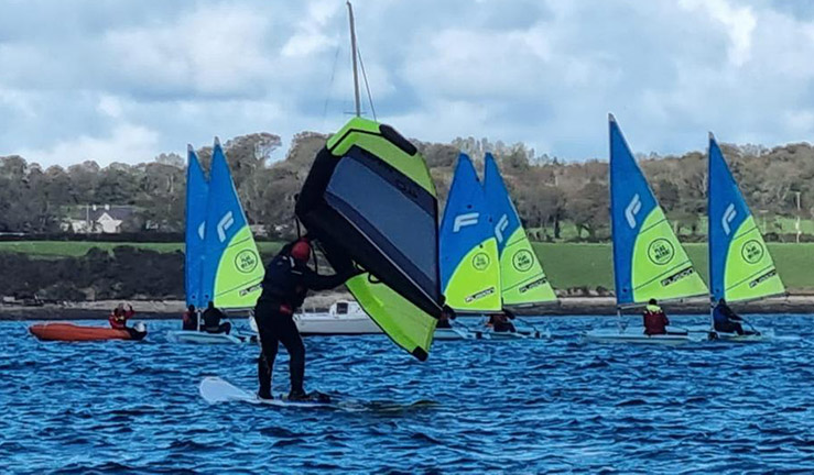 Half a dozen singlehanded dinghies on the water with blue and green sails with a person winging in the foreground at The Big Weekend for clubs, training centres and instructors.