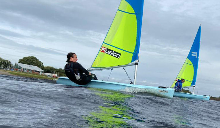 Two people each sailing a Fusion dinghy with blue and green sails against a grey sky in front of clubhouse at Bartley, Birmingham. 