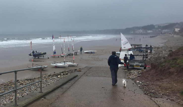 View down the slipway at Filey Sailing Club on a grey day with white waves rolling in onto the beach, with a dog walker in the foreground and youth and junior dinghies starting to put sails up to launch.