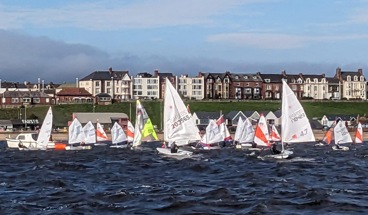 A committee boat on the left with a mixed fleet of youth and junior dinghies starting to line up for a race in the NE & Yorkshire Youth Traveller Series, on the sea at Sunderland Yacht Club on a sunny day with houses on the shoreline.