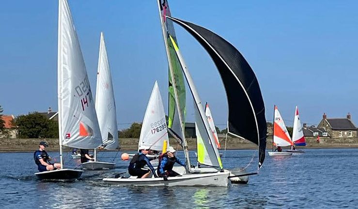 Two juniors in an RS Feva doublehander with a black kite sailing downwind on a blue sky sunny day at Scaling Dam Sailing Club during a North East & Yorkshire Youth Traveller Series event, followed by three ILCA singlehanders and then more junior dinghies.