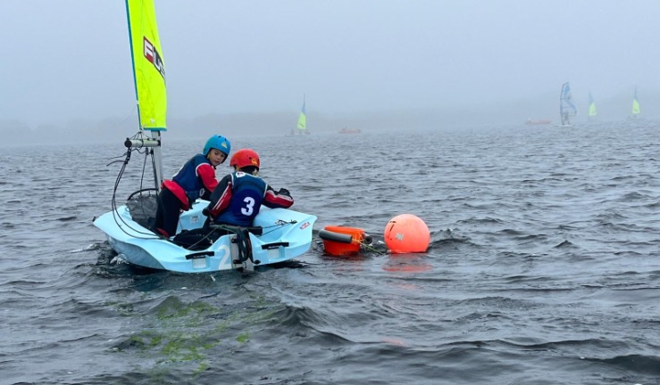 Two children wearing buoyancy aids and helmets, in a dinghy playing a bucket game alongside a buoy