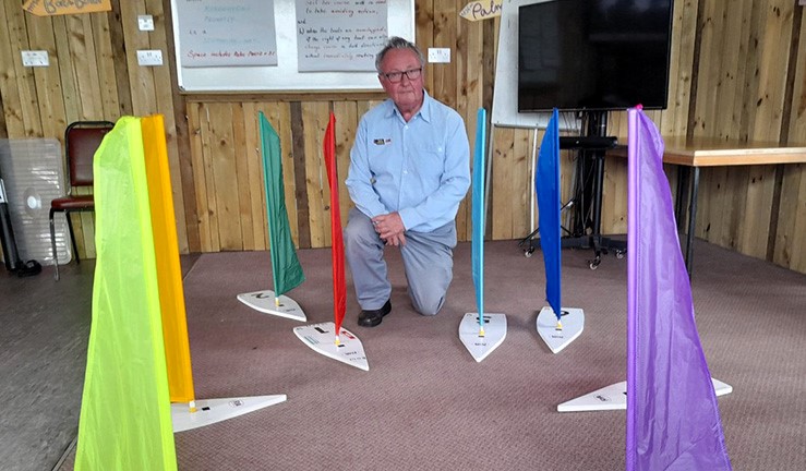 RYA Midlands Regional Rules Advisor Steve Watson kneeling on the floor of a sailing club lounge for a racing rules talk, flanked by 8 large model yachts each with a different colour sail.