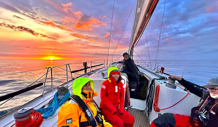 Four crew on offshore kit pictured on a yacht with a colourful sunrise captured before the storms of this year's 50th anniversary Fastnet.