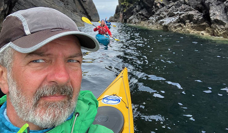 Adventurer and guiding expert Myles Farnbank in a selfie picture on the water in a ravine with two kayakers in the background. 