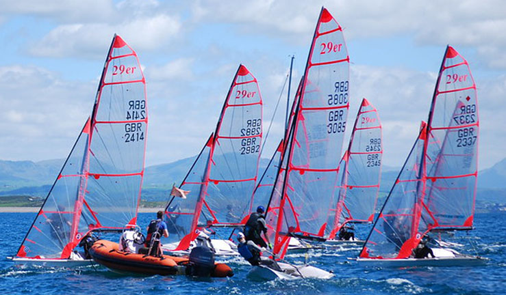 Group of around half a dozen 29ers racing with a RIB in the foreground in Cardigan Bay, Pwllheli, with mountains in the background.