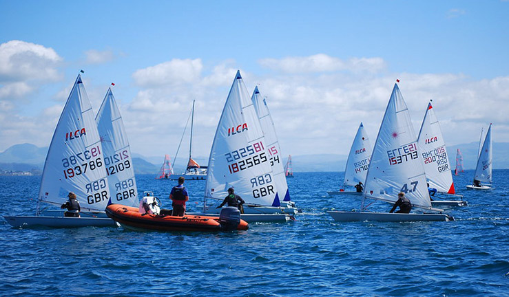Fleet of ILCA dinghy sailors competing at Pwllheli on sunny day with blue skies and blue sea, committee boat and mountains in the background.