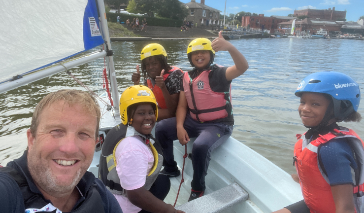 An instructor in a dinghy with four young people wearing helmets and buoyancy aids, all smiling and one giving a thumbs-up