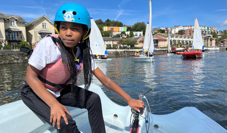 Young girl helming a dinghy, wearing a helmet and buoyancy aid, with other dinghies and dock buildings in the background