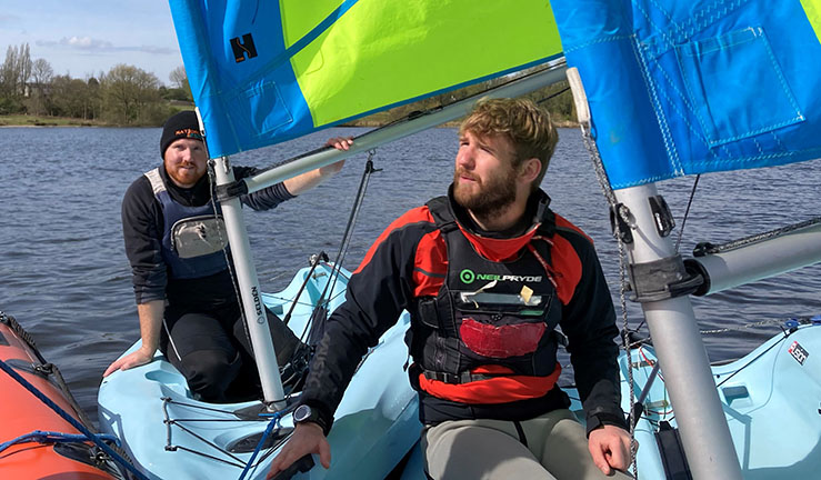 Two instructors sat in blue training dinghies with blue/yellow sails up alongside an orange RIB.