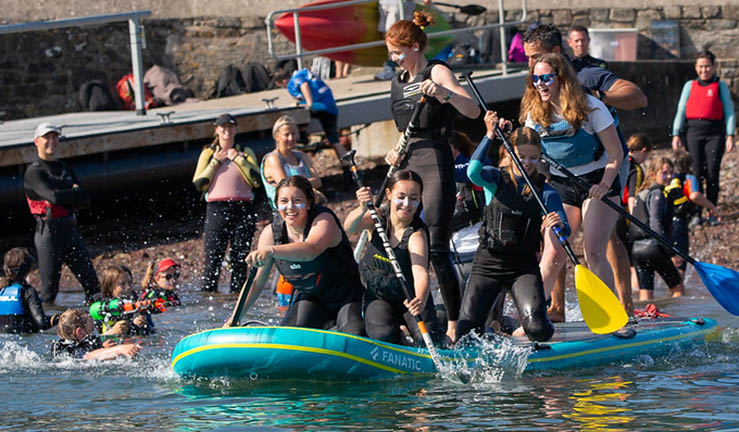 Lots of smiling people having fun on a giant paddleboard at the RYA Cymru Wales OnBoard Festival at Dale.