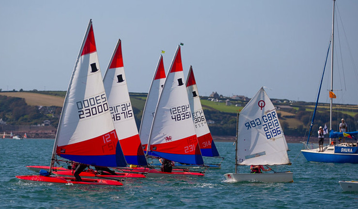 A sunny day with five Topper dinghies with red white and blue sails and an Optimist dinghy crossing the startline near a committee boat for a race at the RYA Cymru Wales OnBoard Festival at Dale.