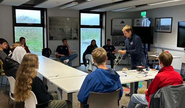British Sailing Team coach Jonny McGovern standing at the front of a classroom demonstrating rope splicing to around half a dozen young people. 