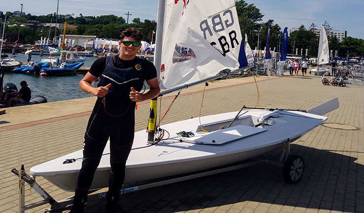 Young sailor standing in front of his dinghy with thumbs up with a GBR sail.