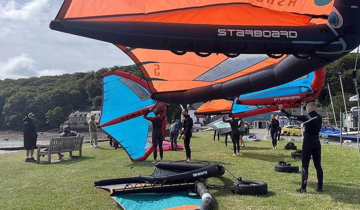 Group of people on shore holding colourful orange and blue wings aloft on a sunny day.