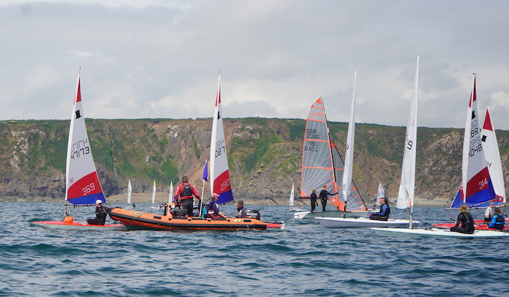 Around half a dozen junior and youth dinghies with colourful sails and an orange RIB on the water with other boats in the background.