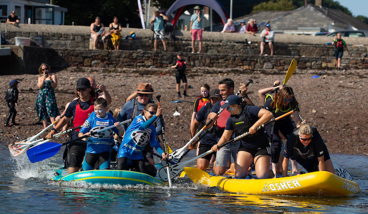 Two groups of people having fun racing away from a beach on two giant paddle boards!