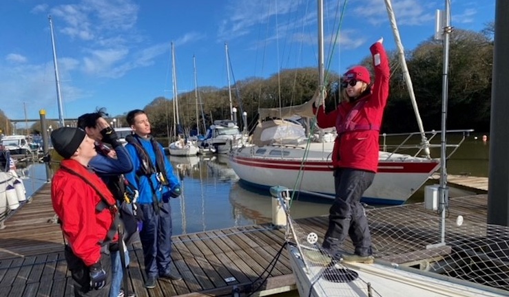 Freya Terry stood on the boat of a yacht in harbour on a sunny day instructing a youth 'pirates' session with three engaged young people listening.
