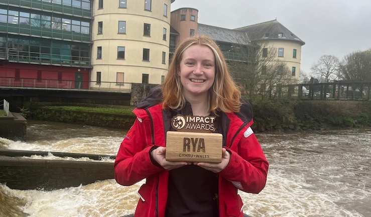 Freya Terry of PPSA Pembrokeshire in a red coat with her wooden RYA Impact Award by the water's edge with buildings in the background.