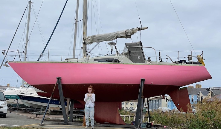 Freya Terry stood in front of the hull of her pink painted yacht Pink Delta which is in a boat yard on shore.