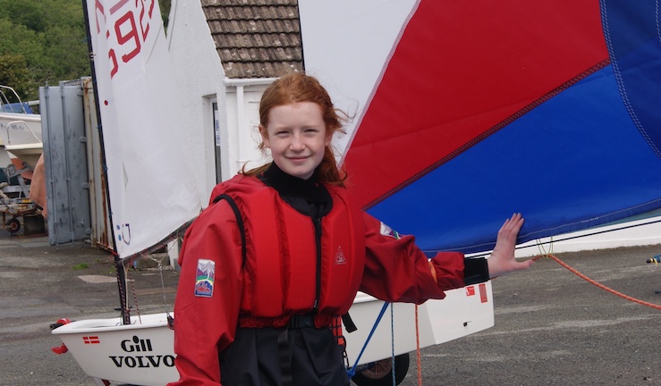 Freya Terry on shore as a young sailor in a red buoyancy aid holding the boom of a red and blue Topper sail with an Optimist dinghy in the background.