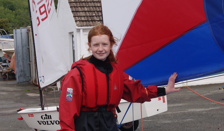 Freya Terry on shore as a young sailor in a red buoyancy aid holding the boom of a red and blue Topper sail with an Optimist dinghy in the background.