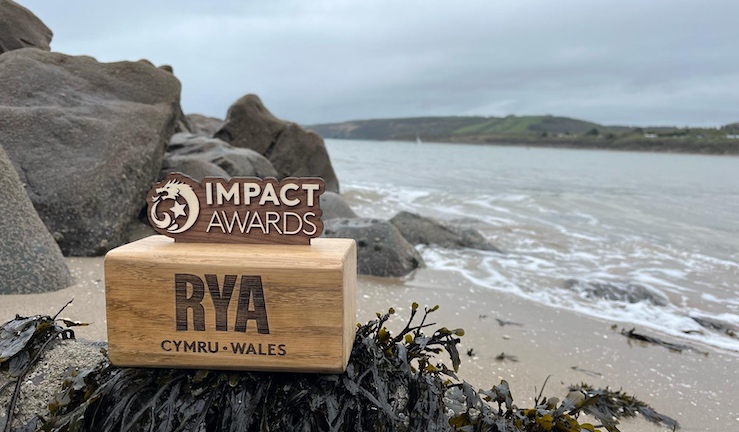 Picture of RYA Cymru Wales Impact Award - wooden dragon logo and words 'IMPACT AWARDS' on a wooden block with RYA Cymru Wales logo, taken outside with view of beach, rocks and coastline in the background.