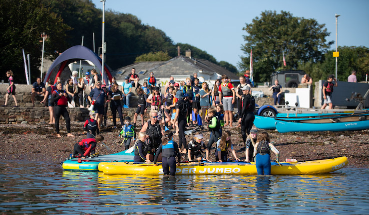 Two giant yellow and and blue paddle boards on the water with lots of people having fun getting on them or on the beach spectating on a sunny day at Dale.