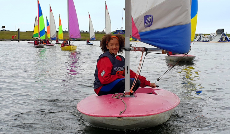 A junior sailor smiling in a red Topper dinghy at Cardiff Sailing Centre with other small dinghies with colourful sails in the background.