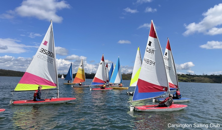 A dozen junior dinghies with multi coloured sails on the water at Carsington on a sunny day with blue skies.