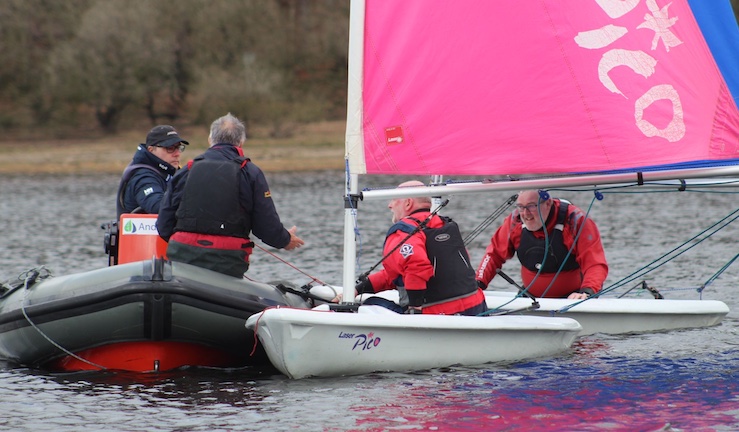 Two instructors in Pico dinghies with pink and blue sails with two instructors in a RIB chatting to them.