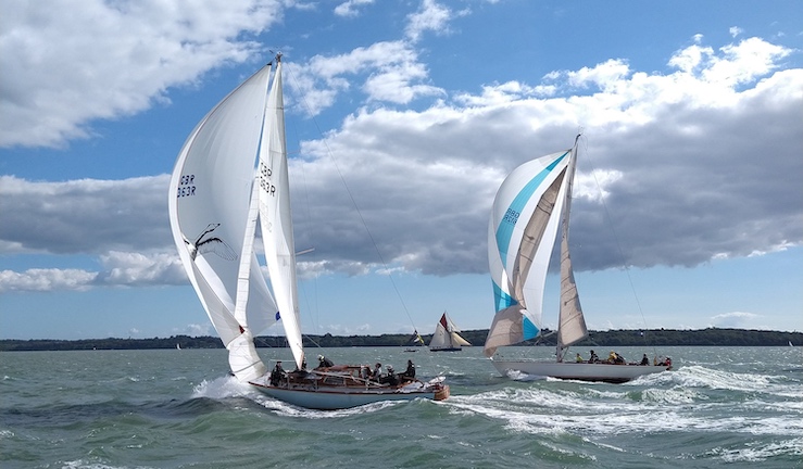 Two classic yachts in the foreground racing with three sails on sunny day with blue skies and white clouds.