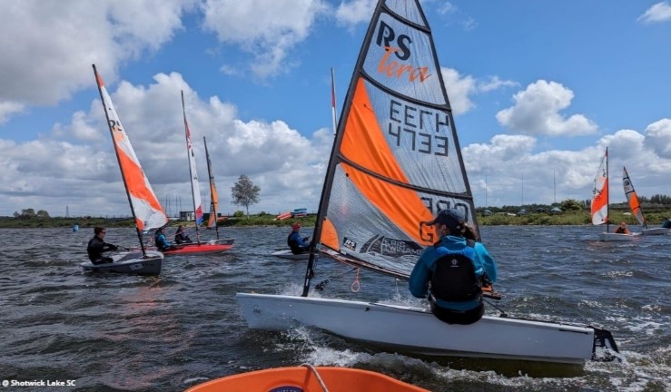 Junior sailing an RS Tera with orange stripe on the sail in the foreground on a sunny day at Shotwick Lake SC for Dragon Series 2024, with half a dozen other junior dinghies in the background.