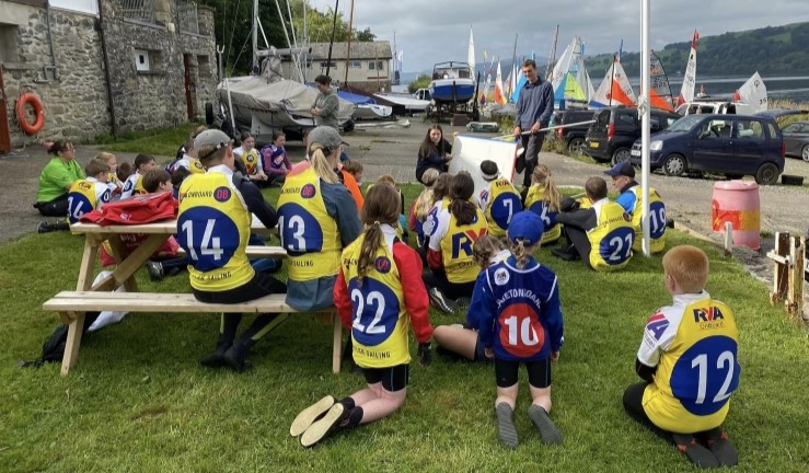 Young sailors on shore wearing yellow OnBoard bibs and listening to a briefing, boats with sails up in the background waiting for launching, at OnBoard Festival, Bala, 2023.