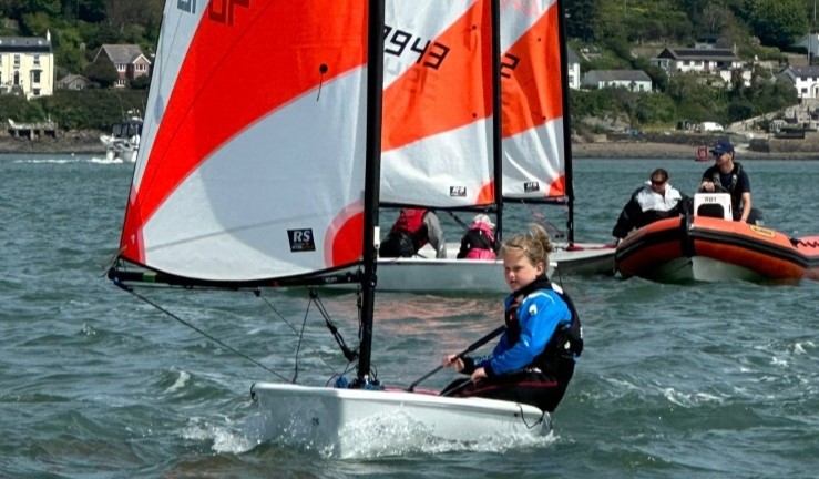 Child sailing an RS Tera dinghy towards the camera with two behind chatting to two coaches on a RIB.