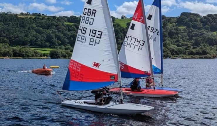 Three Dragon Series sailors helming Topper dinghies with red, white and blue sails, with coach boat following, on a sunny day. 