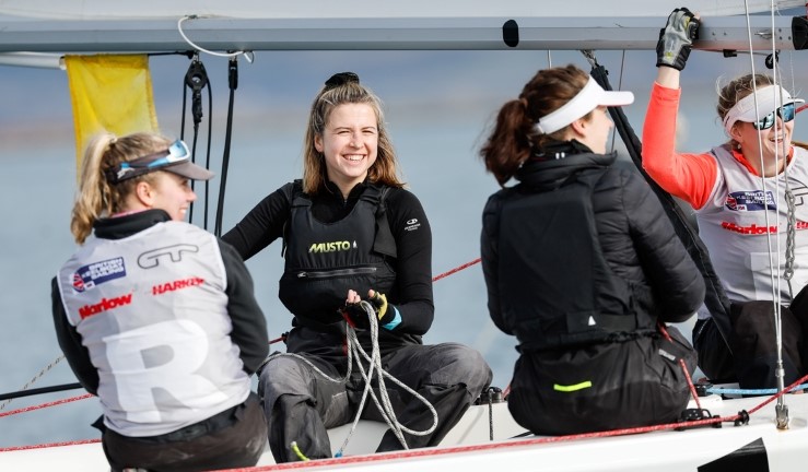 Four girls enjoying sailing a keelboat at an RYA match racing event with one looking directly at the camera with a big smile.