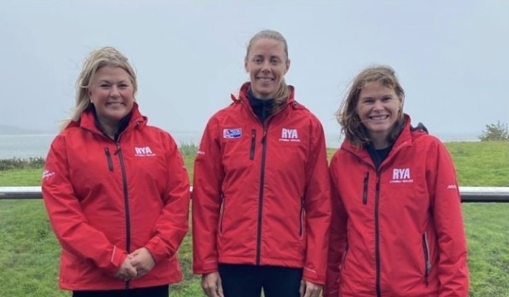 RYA CW Team in branded red jackets and smiling on a grey day, left to right: Chair Sian Reynolds, Performance Manager Sarah McGovern and Club Development Officer Hester Walker.