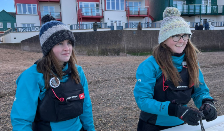 Charlotte left and Anna right, both wearing bobble hats and buoyancy aids, on the beach.