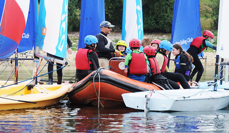 Group of junior sailors in buoyancy aids with dinghies moored around a RIB with an instructor on board.