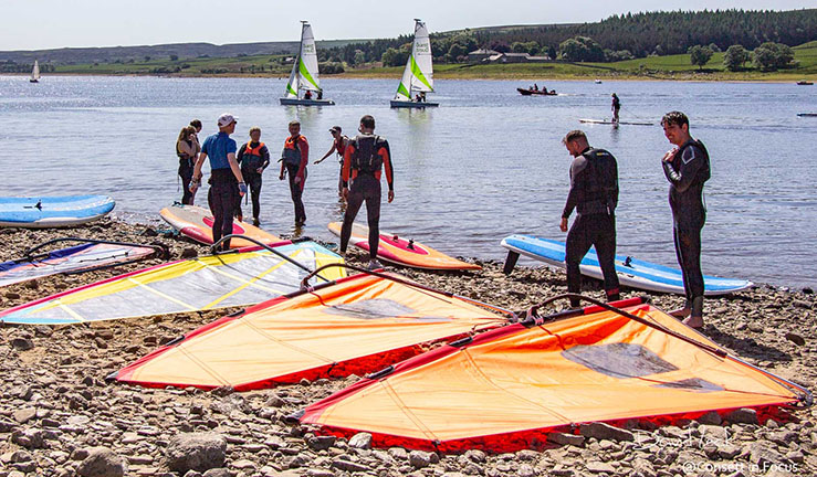 Group of windsurfers on shore with sails on the ground at Derwent Reservoir SC with a couple of sailing dinghies on the water in the background.