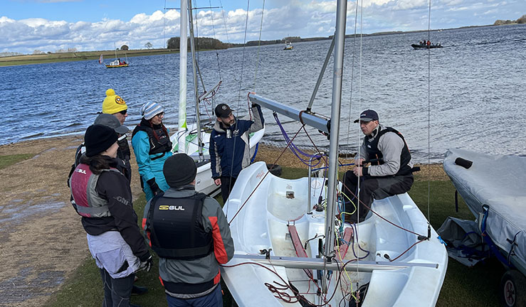 Land drill with trainer in a dinghy on the shore at Rutland Sailing Club and a group gathered around listening at the RYA Midlands Regional Training Day 2023.