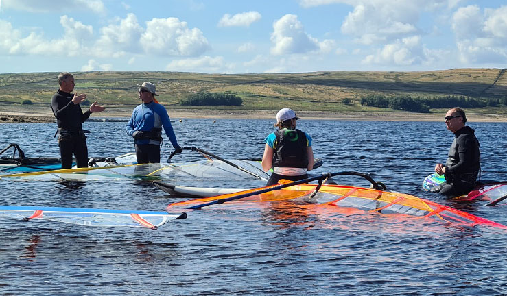 Four people in the water with windsurfing kit on a sunny day with hills in the background and clouds in a blue sky.