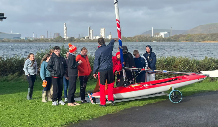 Group of Welsh squad sailors gathered around a Topper dinghy on shore with their coach for a technical talk.