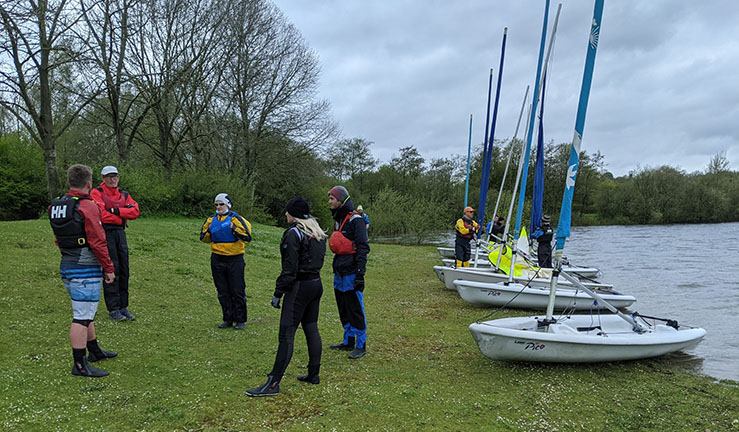 Group of instructors on a course standing in a group on shore in sailing kit with dinghies lined up on the grass alongside a lake waiting to go.