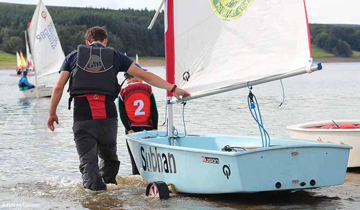 A grown up helping an OnBoard sailor wheel an Optimist dinghy into the water at Derwent Reservoir SC.