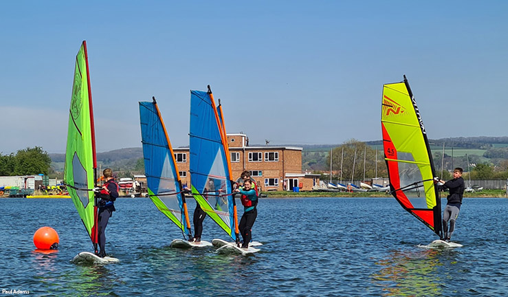 Group of windsurfers sailing towards the camera on a sunny day with Welton Waters Adventure Centre on shore in the background.